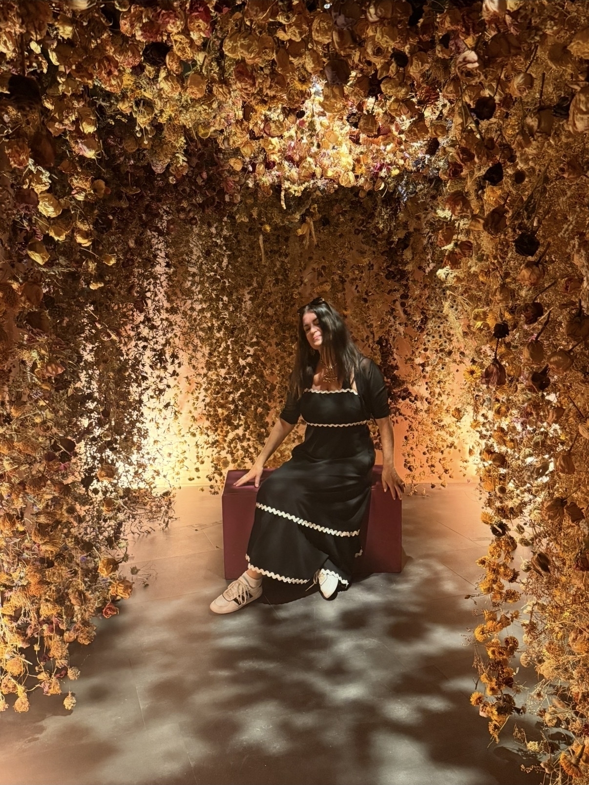 Julia sits on a bench in a black dress with white trim and her dark hair draped around her shoulders, under a golden canopy
